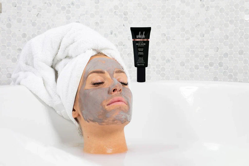 Woman relaxing in a towel with a gray mud mask applied and a Whish beauty mud mask tube placed beside her.