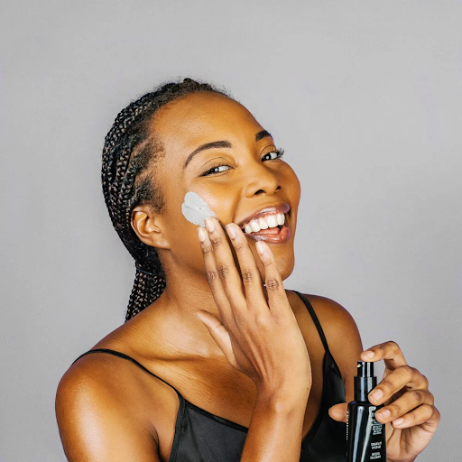 A woman with braided hair applies a gray mud mask from Whish to her cheek, holding a black pump bottle.
