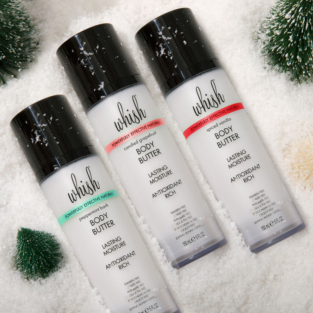 Three Whish Beauty body butter bottles displayed in winter snow with small evergreen trees.