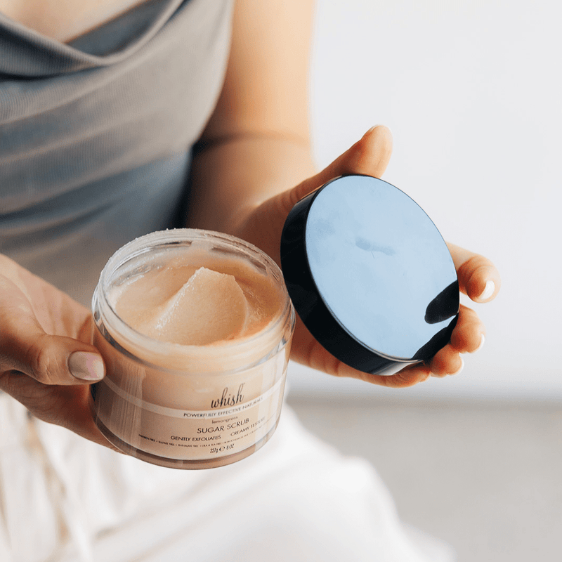 A person holds an open jar of Whish Sugar Scrub, showing its creamy texture.