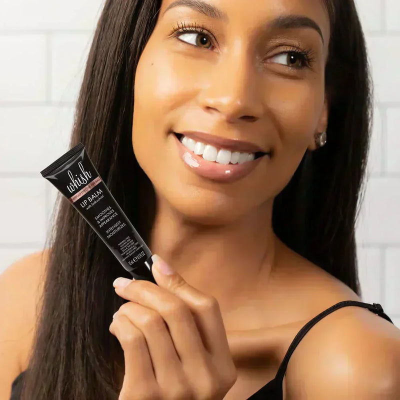 Woman smiling while holding Whish Beauty bakuchiol lip balm in a bright white-tiled bathroom.