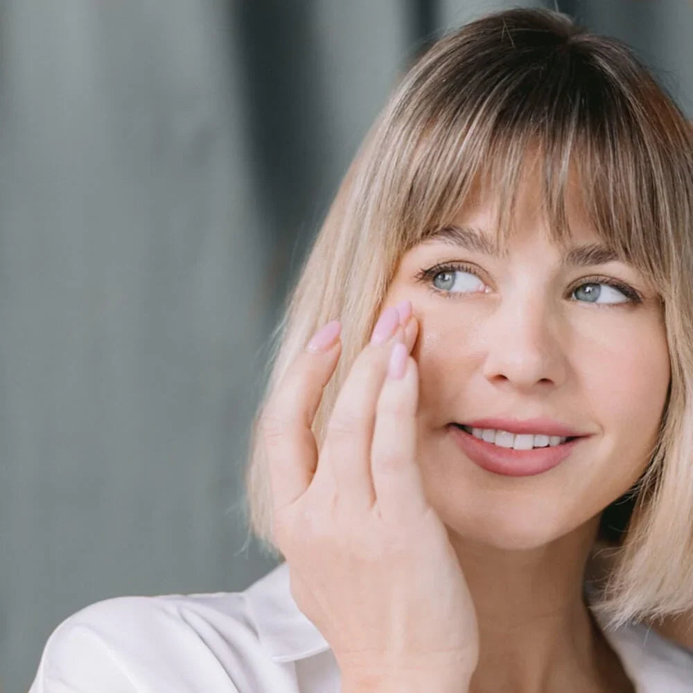 Blonde woman smiling while applying Whish Vitamin C+E Serum near the eye area as part of a gentle skincare routine for dry, sensitive skin.