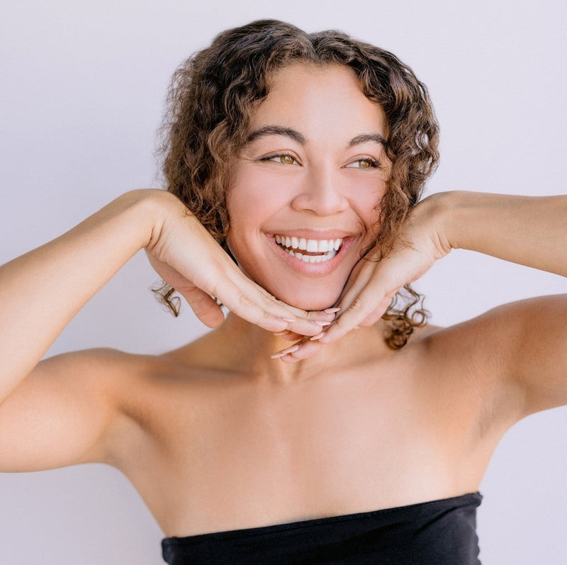 Woman with curly hair wearing a black top, raising her arms near her face against a neutral background.